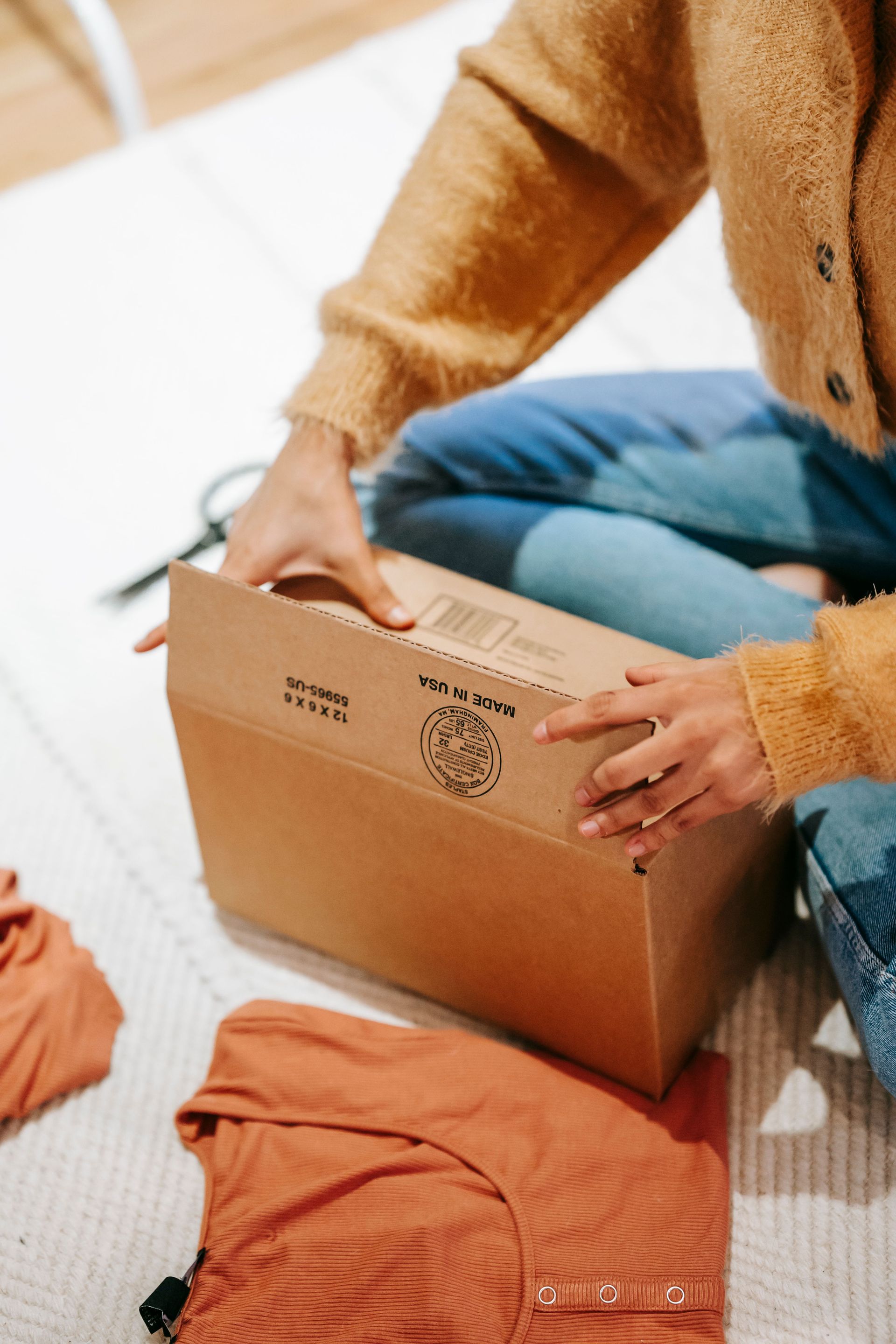 From above of crop anonymous female opening carton box with cotton clothes on floor