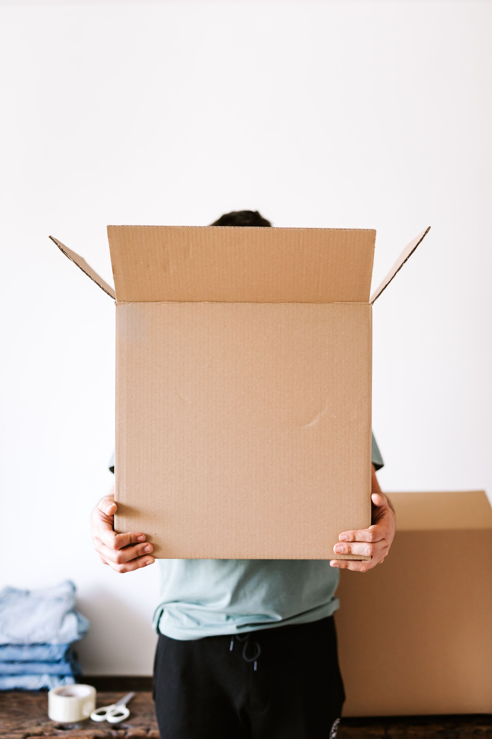 Anonymous man carrying opened carton box before packing belongings and relocation while standing against white wall and stacked clothes